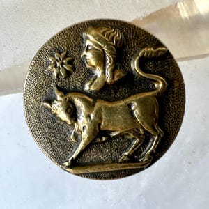 May include: A round, antique brass-colored button with a textured background. The button features a relief design of a bull, a woman's profile, and a small floral element. The bull is depicted in a walking pose.