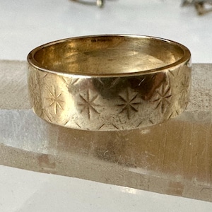 May include: A gold-colored ring with a wide band. The ring is decorated with a repeating pattern of etched starburst designs. The ring is displayed on a clear, rectangular surface.