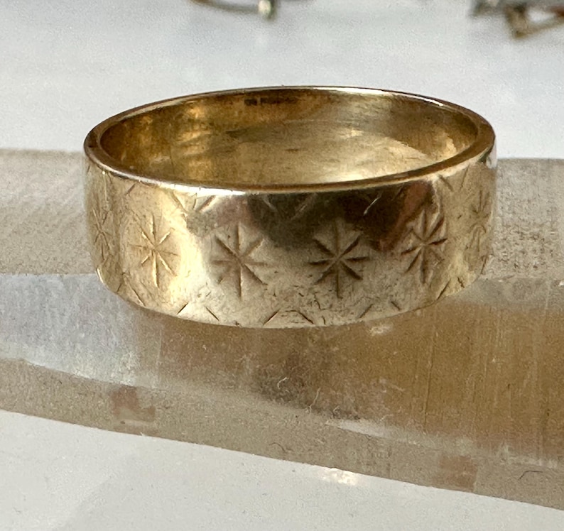 May include: A gold-colored ring with a wide band. The ring is decorated with a repeating pattern of etched starburst designs. The ring is displayed on a clear, rectangular surface.