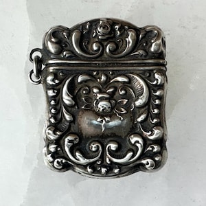 May include: Ornate silver match safe with a hinged lid. The rectangular case features detailed floral and scrollwork designs. A small loop is attached for a chain. The antique piece has a tarnished finish.