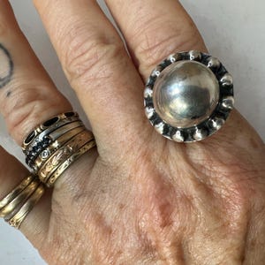 May include: A large, silver ring with a rounded central design and a beaded edge. Other rings in gold and black are also visible on the hand. A tattoo is present on the index finger. The image showcases a collection of jewellery.