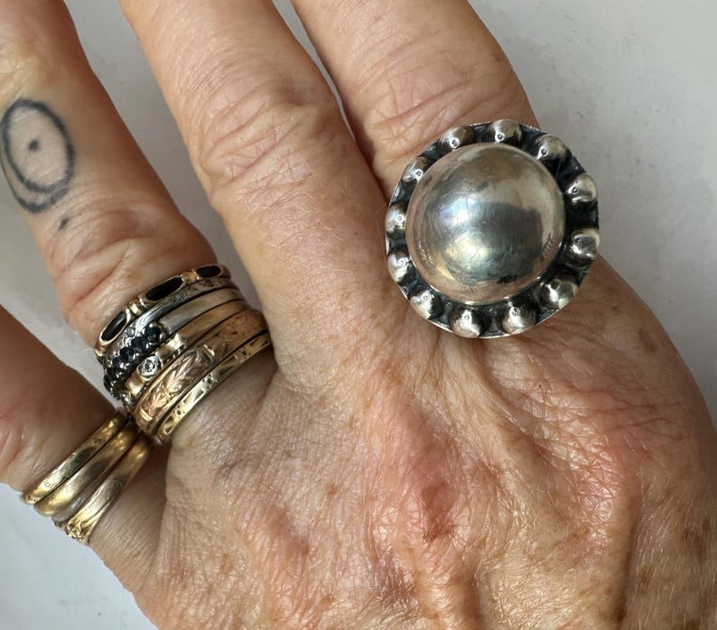 May include: A large, silver ring with a rounded central design and a beaded edge. Other rings in gold and black are also visible on the hand. A tattoo is present on the index finger. The image showcases a collection of jewellery.