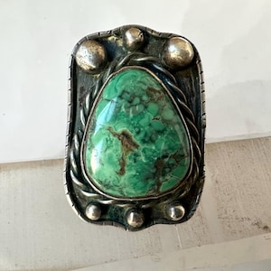 May include: Vintage silver-tone ring featuring a large, teardrop-shaped turquoise stone. The stone is a vibrant green with brown veining. The ring has a rectangular frame with decorative silver accents and three silver balls at the top.