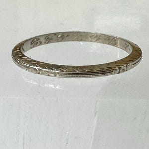 May include: A silver-colored ring with an etched design. The ring has a narrow band with a repeating geometric pattern around the exterior. The ring appears to be vintage or antique, with some wear visible on the surface.
