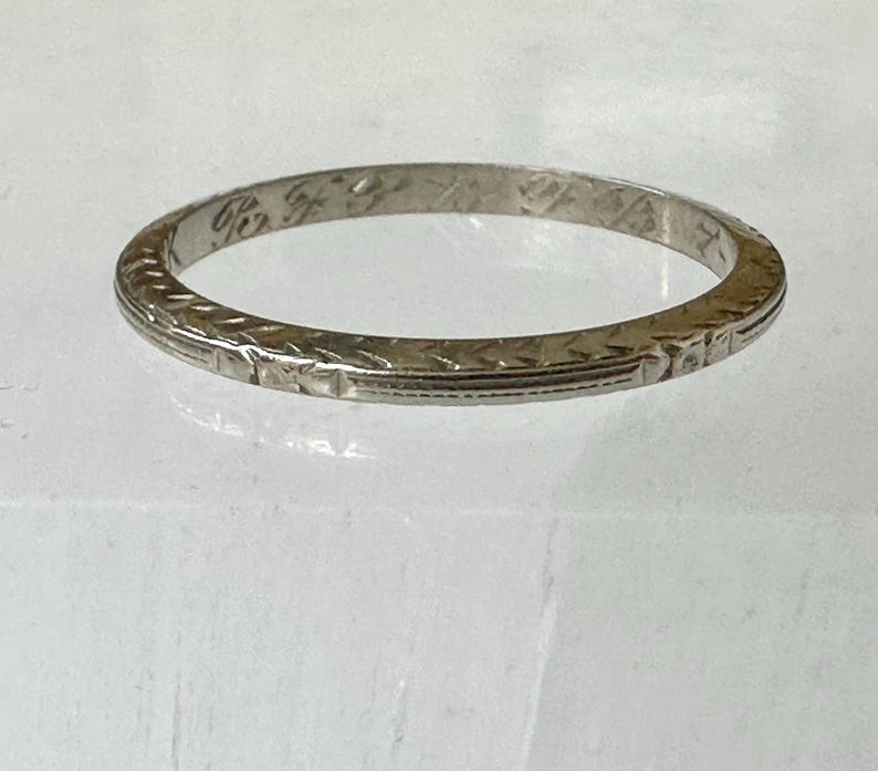 May include: A silver-colored ring with an etched design. The ring has a narrow band with a repeating geometric pattern around the exterior. The ring appears to be vintage or antique, with some wear visible on the surface.