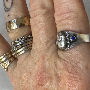 May include: A hand displays several rings, including a gold band with starburst engravings, and a silver ring with a scarab beetle design and a blue gemstone. Other rings include floral and twisted metal designs.