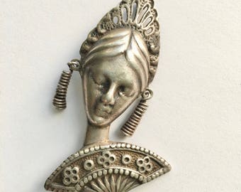 May include: Silver brooch featuring a woman's face with a crown and a fan. The woman's face is detailed with a closed mouth and eyes. The fan is open and has a decorative design.