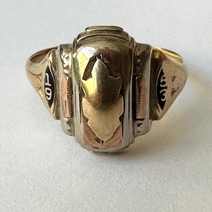 May include: A gold class ring with a black enamel inscription of '19' on the side. The ring has a raised, gold-toned design in the center.
