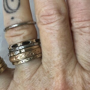 May include: A hand displays a collection of gold and silver rings. The rings vary in design, including bands with black enamel, floral patterns, and small gemstones. One ring is a simple, thin silver band. The rings are stacked on multiple fingers.