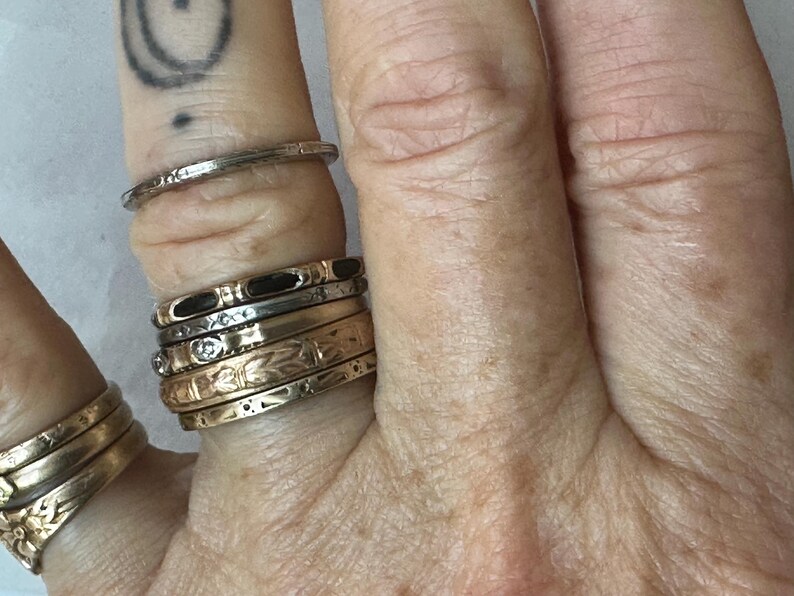 May include: A hand displays a collection of gold and silver rings. The rings vary in design, including bands with black enamel, floral patterns, and small gemstones. One ring is a simple, thin silver band. The rings are stacked on multiple fingers.