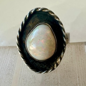 May include: A silver ring featuring an oval, iridescent opal stone. The stone is set within a dark, textured bezel, surrounded by a twisted rope design. The ring's band is a simple, polished silver.