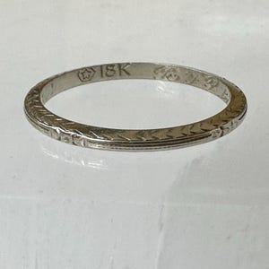 May include: A thin, silver-colored ring with an engraved pattern. The ring is marked with "18K" and other symbols. The band features a repeating geometric design. The ring is likely made of gold.