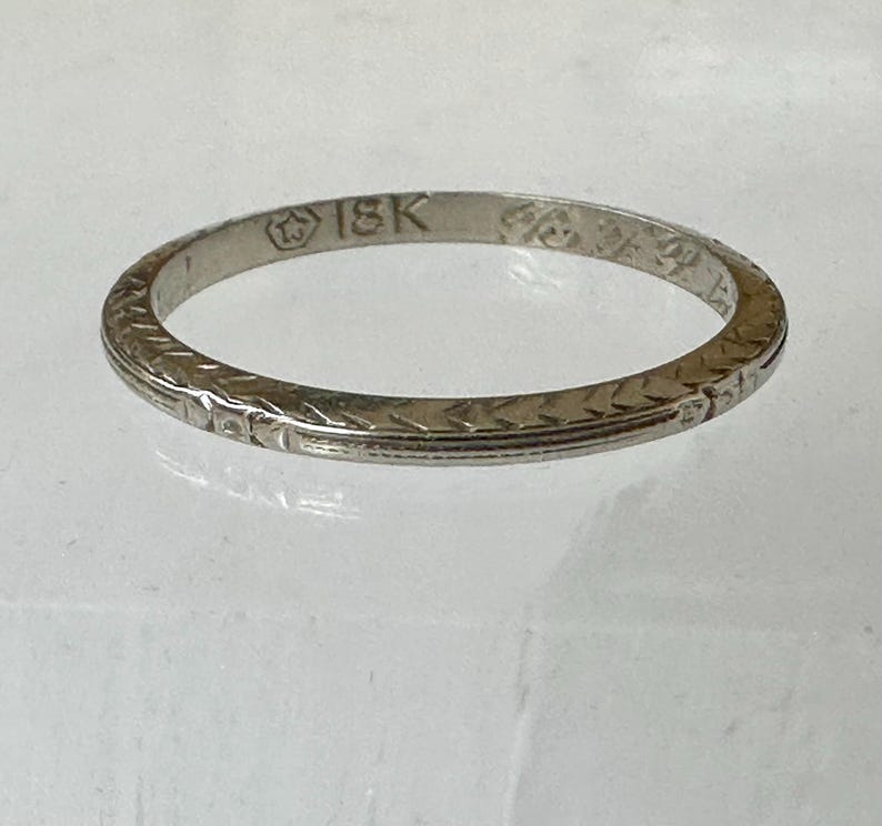 May include: A thin, silver-colored ring with an engraved pattern. The ring is marked with "18K" and other symbols. The band features a repeating geometric design. The ring is likely made of gold.