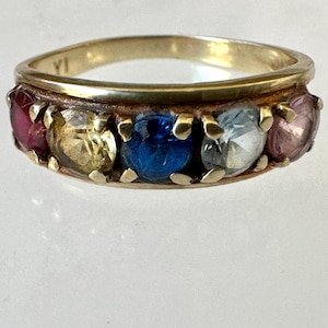 May include: A gold-toned ring featuring a row of colorful gemstones. The gemstones include red, yellow, blue, clear, and purple stones. The ring has a classic design, with the gemstones set in a band.