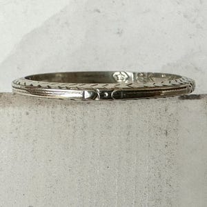 May include: A silver-toned ring with intricate etched patterns. The ring features a repeating geometric design along the band's edge and a central rectangular detail. The ring is set against a neutral background, highlighting its detailed craftsmanship.