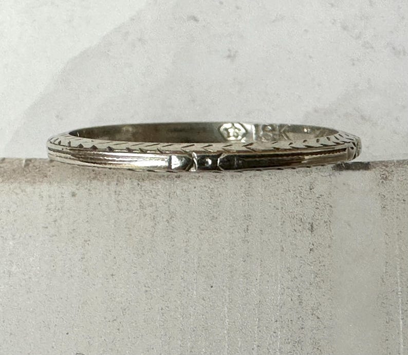 May include: A silver-toned ring with intricate etched patterns. The ring features a repeating geometric design along the band's edge and a central rectangular detail. The ring is set against a neutral background, highlighting its detailed craftsmanship.