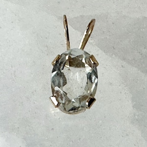May include: A clear, oval-cut gemstone pendant set in a gold-toned metal prong setting. The faceted stone displays a transparent, colorless appearance, secured by four small prongs at the base and topped with a gold-toned metal bail for hanging.