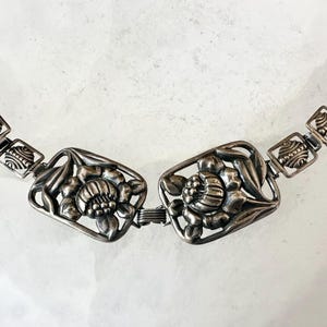 May include: A silver-toned metal belt featuring a floral design. The belt has two large rectangular centerpieces with detailed flower motifs, connected by a clasp. The chain links are square with decorative patterns.