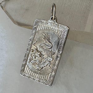 May include: Silver rectangular pendant featuring a detailed dragon design. The pendant has a loop at the top for a chain and a decorative border. The dragon is the focal point, with intricate scales and a dynamic pose. The pendant is likely made of sterling silver.