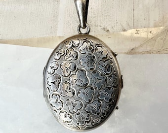Op de afbeelding: Ovale zilveren hanger met een bloemmotief. De hanger heeft een lus aan de bovenkant voor een ketting en een sluiting aan de zijkant. Het oppervlak van de hanger is gegraveerd met een herhalend ontwerp van gestileerde bloemen.