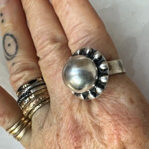 May include: A silver ring featuring a large, polished, spherical centre stone, surrounded by a beaded border. The ring is on a hand, alongside other rings of various designs and a small tattoo.