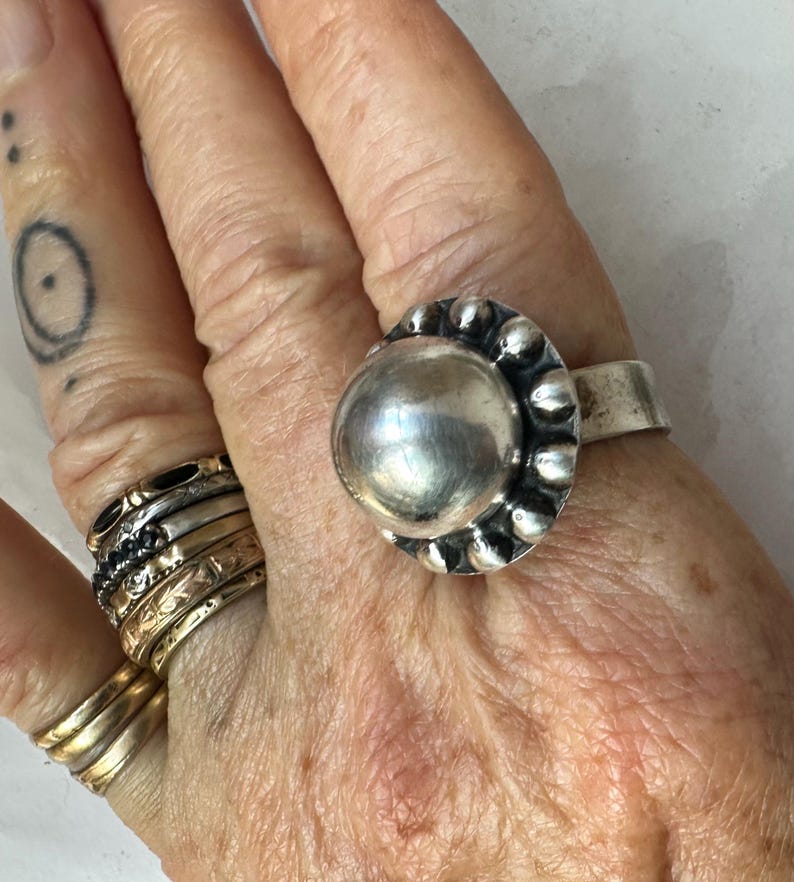May include: A silver ring featuring a large, polished, spherical centre stone, surrounded by a beaded border. The ring is on a hand, alongside other rings of various designs and a small tattoo.