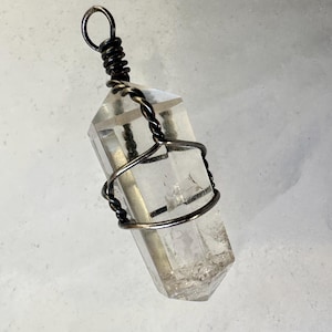 May include: A clear, hexagonal quartz crystal pendant wrapped in dark, oxidized metal wire. The wire forms a spiral cage around the crystal body and terminates in a circular loop at the top for suspension, showcasing natural internal inclusions.