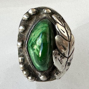 May include: Oval-shaped silver ring with a vibrant green malachite stone center. The ring features a leaf design on one side and a beaded border. A unique piece of jewelry.