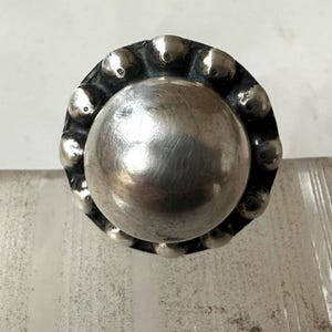 May include: A silver ring featuring a large, polished central sphere. The ring's band is decorated with small, raised circular details around the central sphere. The ring has a vintage or antique appearance.