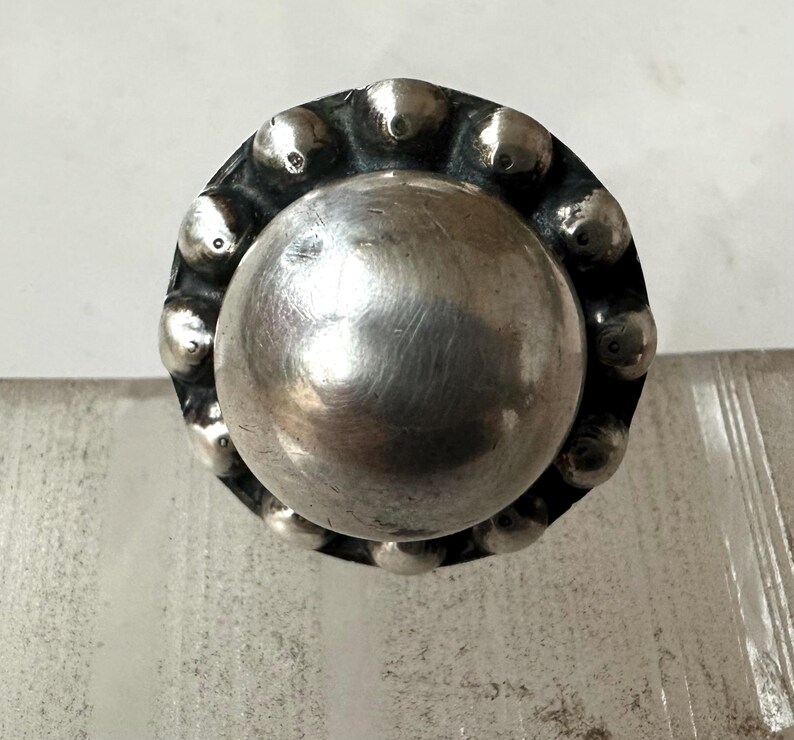 May include: A silver ring featuring a large, polished central sphere. The ring's band is decorated with small, raised circular details around the central sphere. The ring has a vintage or antique appearance.