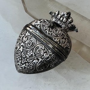 May include: A silver-toned, heart-shaped container with intricate floral and scrollwork engravings. A small crown adorns the top. The container is likely a decorative box or pendant, showcasing detailed craftsmanship.