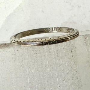May include: A thin, silver-colored ring with an engraved pattern. The ring is marked with "18K" and has a delicate, textured design. The ring is likely made of gold and is a piece of jewelry.