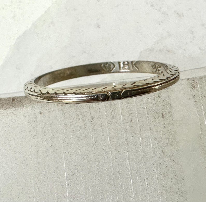 May include: A thin, silver-colored ring with an engraved pattern. The ring is marked with "18K" and has a delicate, textured design. The ring is likely made of gold and is a piece of jewelry.