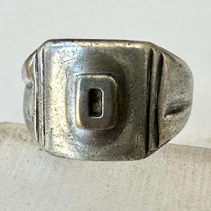 May include: A silver-toned ring with a rectangular face. The face features a recessed square with the letter "D" in the center. Vertical lines are etched on either side of the face. The ring has a worn, vintage appearance.