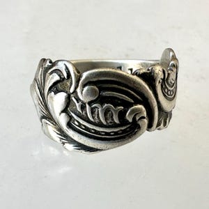 May include: A silver-toned ring with ornate, swirling floral and scrollwork designs. The ring features a central oval element with a decorative pattern and is likely made of sterling silver or a similar metal.