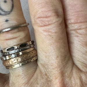 May include: Close-up of a hand adorned with multiple rings. The rings vary in style, including textured bands, bands with small diamonds, and bands with intricate patterns. The rings are in gold and silver tones.