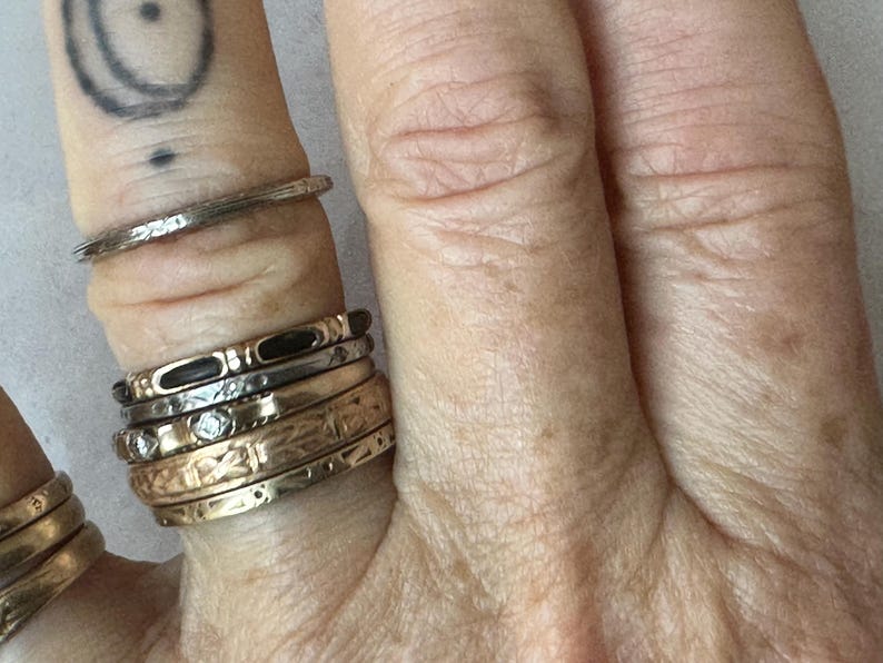 May include: Close-up of a hand adorned with multiple rings. The rings vary in style, including textured bands, bands with small diamonds, and bands with intricate patterns. The rings are in gold and silver tones.