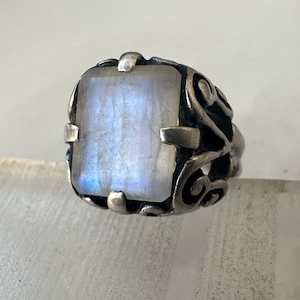 May include: A silver ring with a large, rectangular, iridescent moonstone. The moonstone has a soft, glowing appearance with blue and white hues. The ring has an ornate, swirling design around the stone.