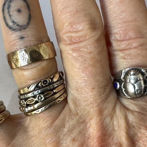 May include: A collection of rings in various styles and metals. The rings include a gold band with starburst engravings, a silver ring with a scarab beetle design and blue accents, and several stacked gold and silver bands with floral and geometric patterns.
