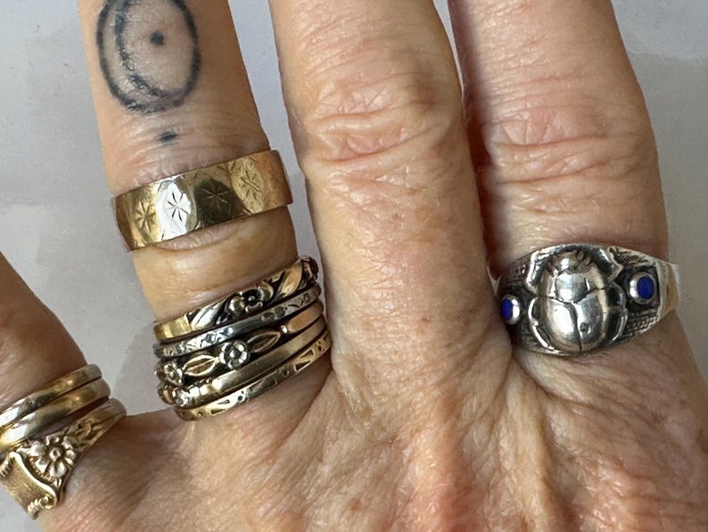 May include: A collection of rings in various styles and metals. The rings include a gold band with starburst engravings, a silver ring with a scarab beetle design and blue accents, and several stacked gold and silver bands with floral and geometric patterns.