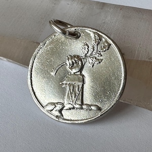 May include: Silver charm featuring a stylized hand holding a branch with leaves. The charm is round and has a smooth, metallic finish.