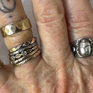May include: A hand displays a collection of rings in various styles and metals. The rings include a wide gold band with starburst engravings, several delicate floral bands, and a silver ring featuring a scarab beetle design with blue accents.