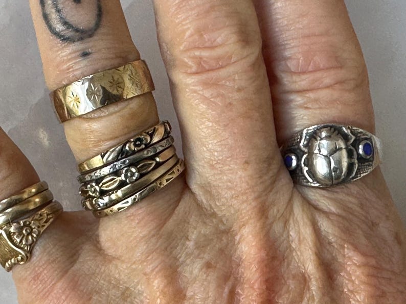 May include: A hand displays a collection of rings in various styles and metals. The rings include a wide gold band with starburst engravings, several delicate floral bands, and a silver ring featuring a scarab beetle design with blue accents.