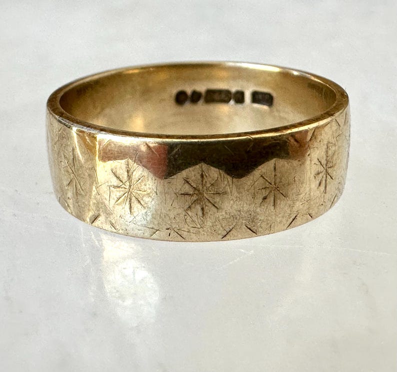 May include: A gold-colored ring with a wide band. The ring's surface is etched with a repeating pattern of starburst designs. The ring has a slightly worn appearance, suggesting age or use.