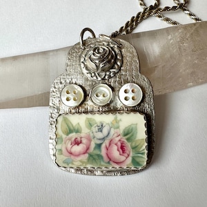 May include: A silver pendant necklace with a floral design. The pendant features a silver rose, three white pearl buttons, and a rectangular porcelain tile with a floral pattern. The necklace is on a silver chain.