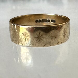 May include: A gold-colored ring with a wide band. The ring is engraved with a repeating pattern of starburst designs. The ring has markings inside the band. The ring is displayed on a light-colored surface.