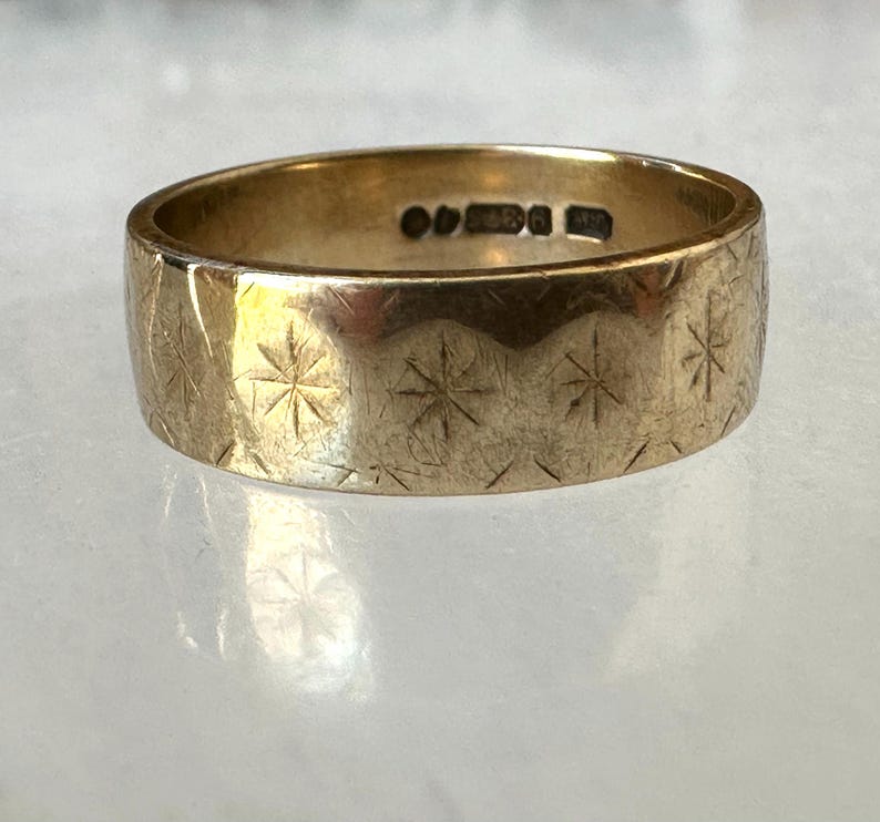 May include: A gold-colored ring with a wide band. The ring is engraved with a repeating pattern of starburst designs. The ring has markings inside the band. The ring is displayed on a light-colored surface.