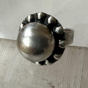May include: A silver ring featuring a large, polished, spherical central element. The bezel is decorated with small, rounded, textured embellishments. The band is a plain, thick, silver band. The ring is shown on a clear, textured surface.