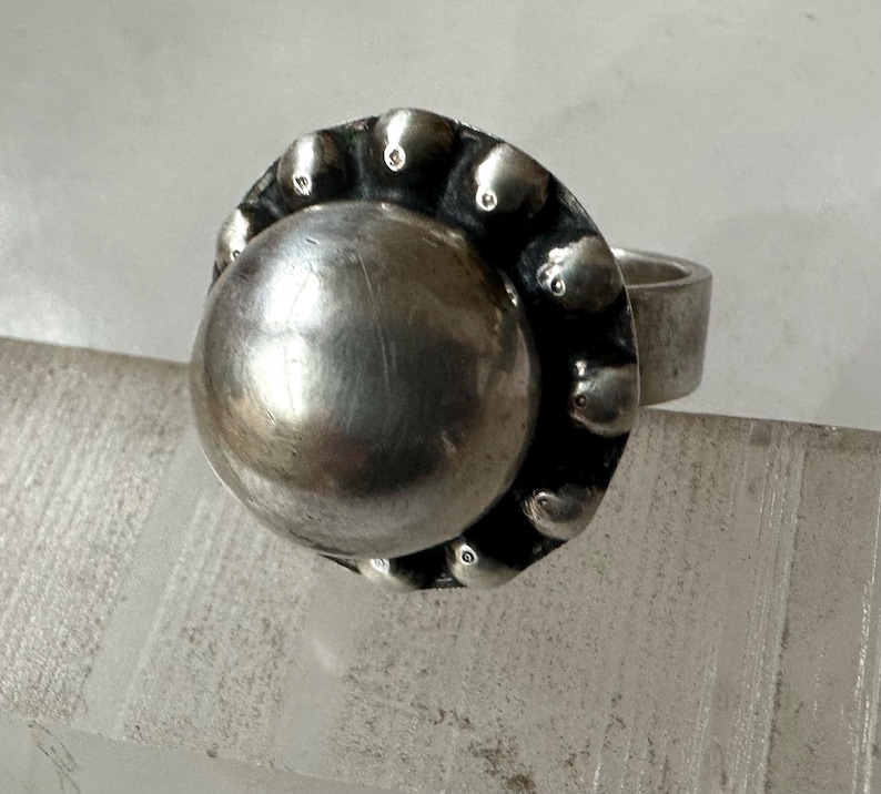 May include: A silver ring featuring a large, polished, spherical central element. The bezel is decorated with small, rounded, textured embellishments. The band is a plain, thick, silver band. The ring is shown on a clear, textured surface.