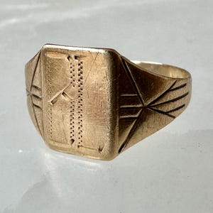 May include: A gold-toned signet ring featuring a rectangular face with an engraved vertical line pattern and decorative etched lines extending along the sides of the band. The metal surface shows visible signs of wear and fine surface scratches.